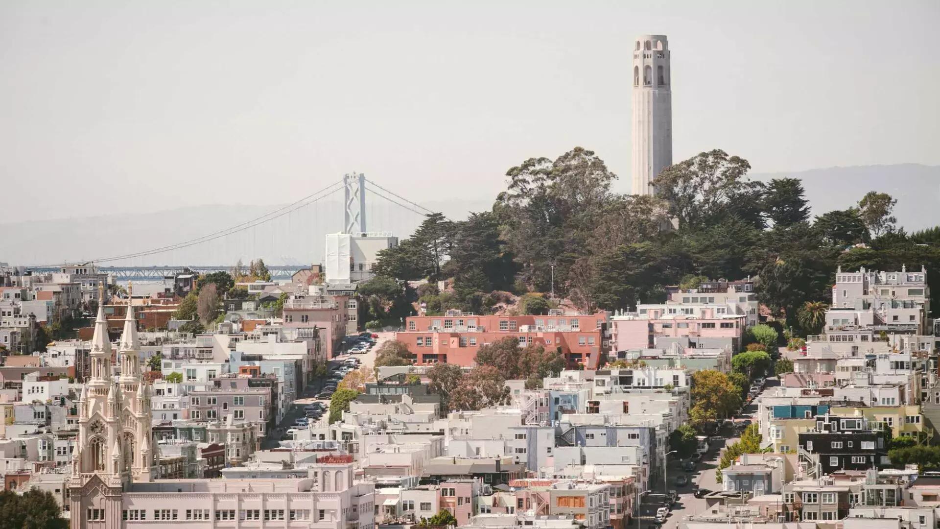 San Francisco's Coit Tower is pictured with the Bay Bridge in the background and a hill covered in houses in the foreground.