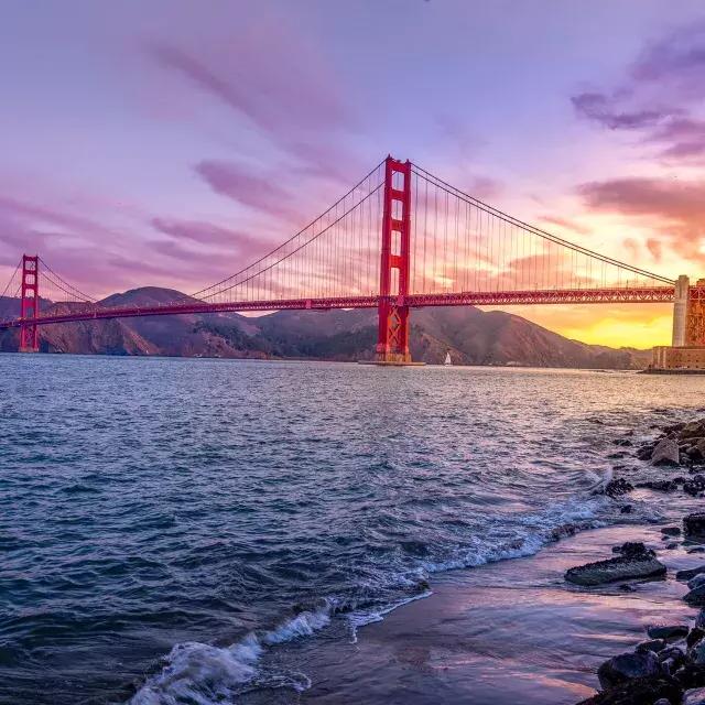 The Golden Gate Bridge at sunset with a multicolored sky and the San Francisco Bay in the foreground.